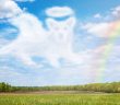A cloud shaped like a dog with a halo floats in a bright blue sky above a green field, with a soft rainbow arching through the sky.
