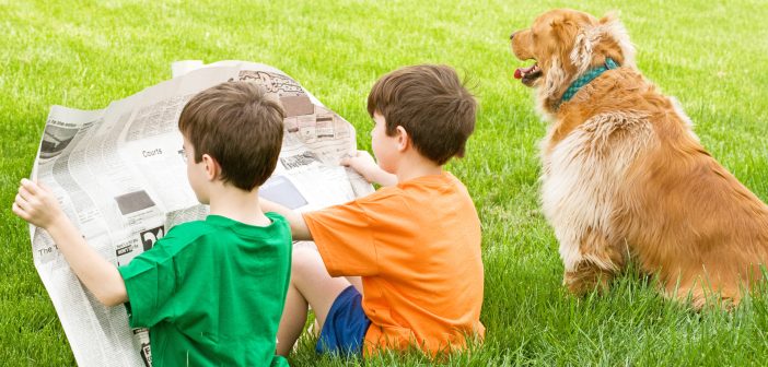 two boys sharing a newspaper and a dog all sitting outside in the grass
